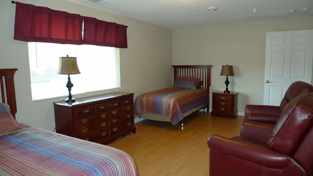 A bright bedroom with two twin beds, wooden dressers, bedside lamps, and two leather chairs.