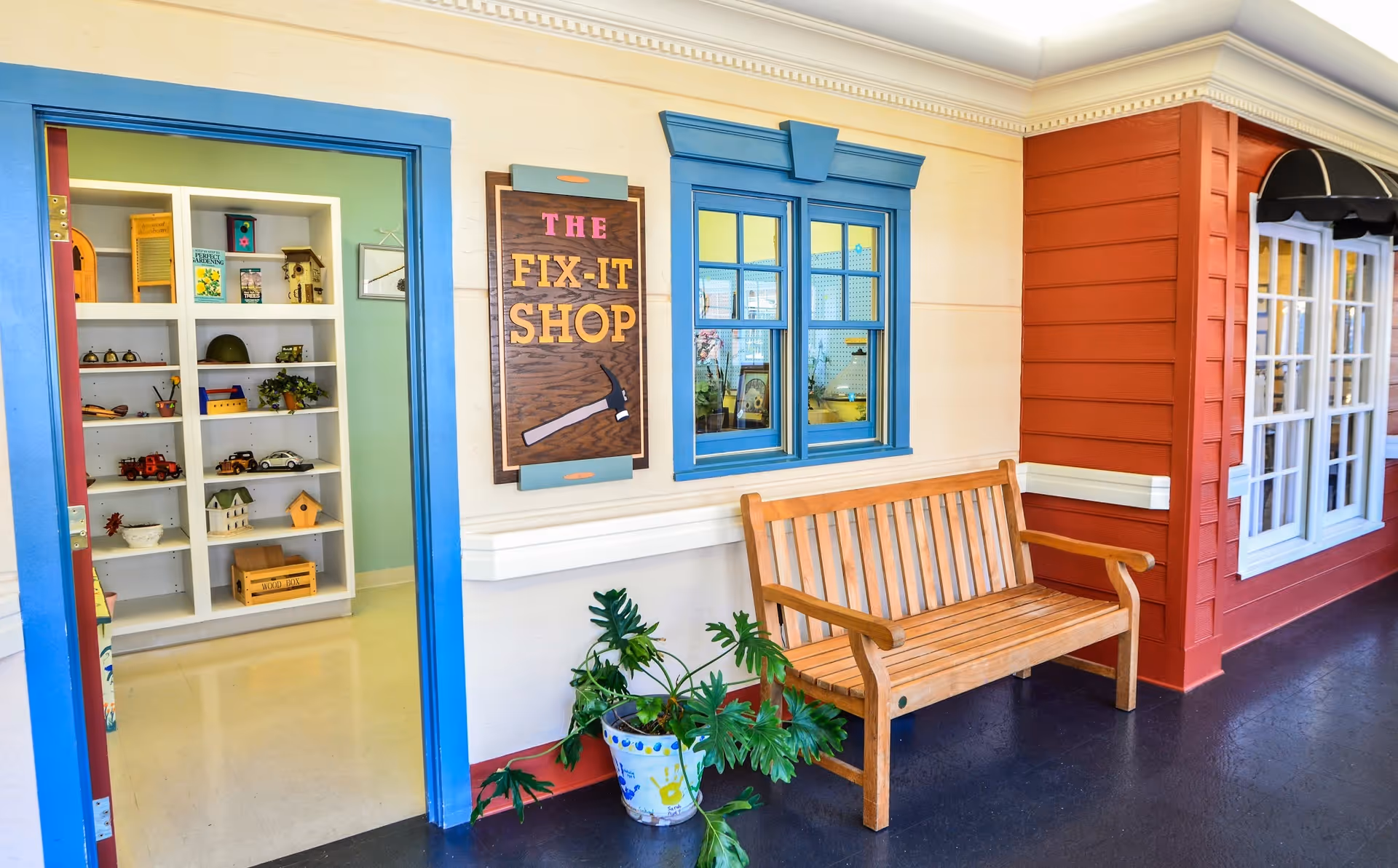 A hallway with a wooden bench and a potted plant next to a blue-framed window and door. The door leads to a room with shelves holding various small items and decorations. A sign on the wall reads 'The Fix-It Shop' with an image of a hammer.
