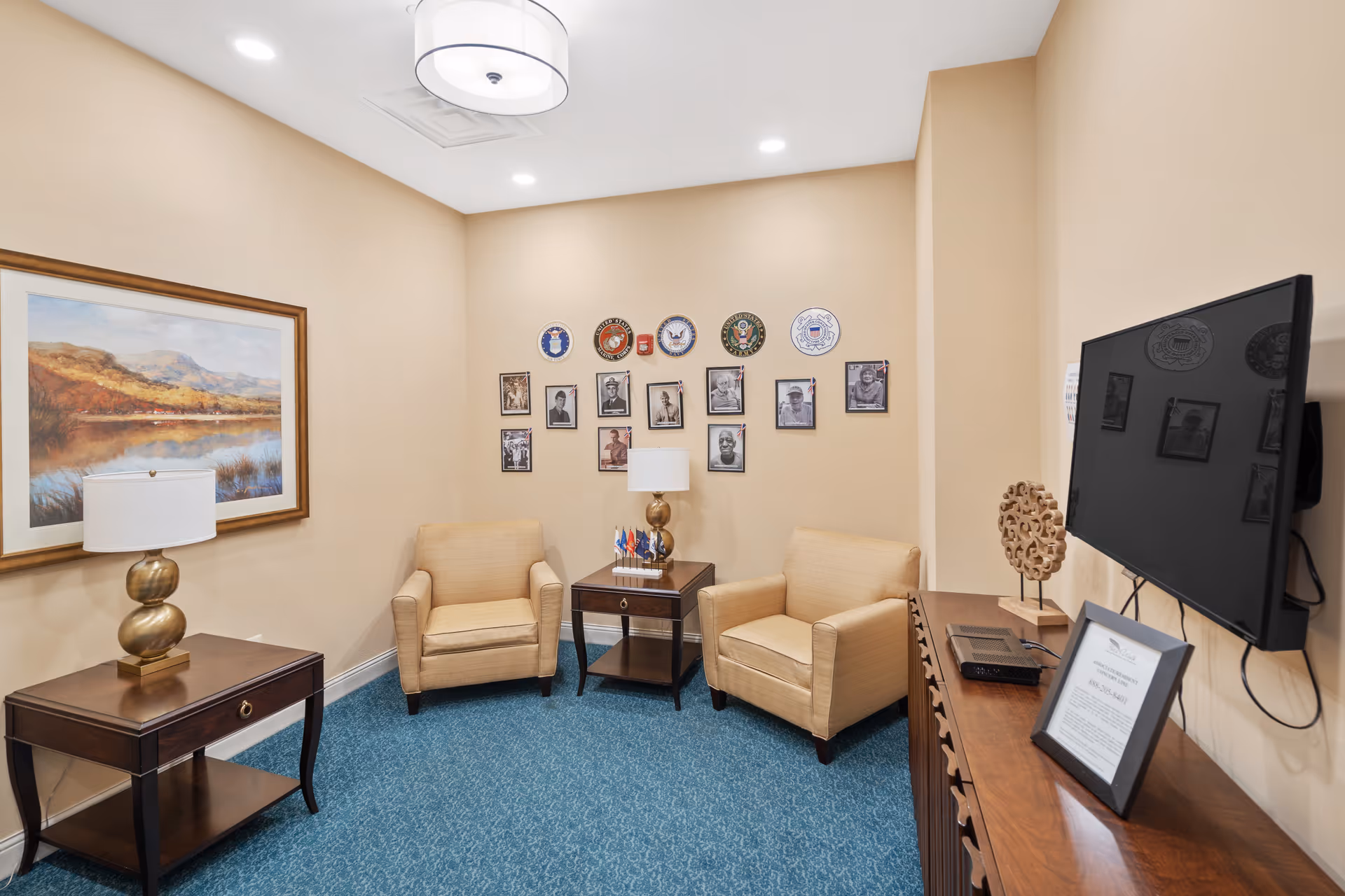 Small sitting room with two beige armchairs, side tables, a wall-mounted TV, and framed photos and plaques on a beige wall.
