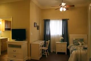 A cozy bedroom with a single bed on the right side, a nightstand with a flower vase next to the bed, a window with dark curtains in the center, a small desk and chair against the left wall, and a dresser with a flat-screen TV on top. The room has warm yellow walls and a ceiling fan with lights.