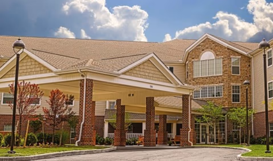Front entrance of a multi-story senior living building with a covered porte-cochère supported by brick columns and landscaped grounds under a partly cloudy sky.