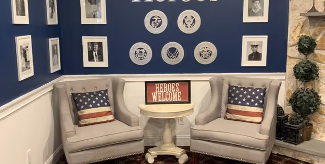 Small seating area with two upholstered armchairs, American flag pillows, a round table with a 'HEROES WELCOME' sign, and framed photos and service emblems on a blue wall.