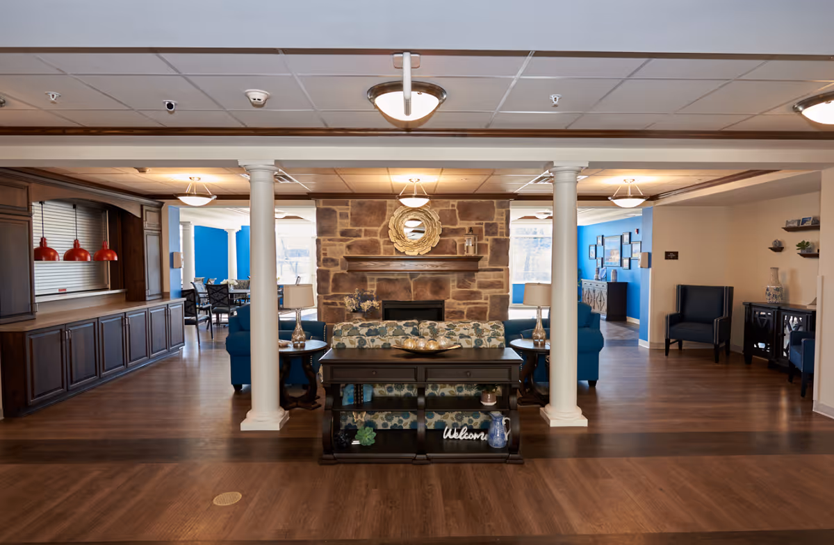 A cozy common area in a senior living facility featuring a stone fireplace with a decorative mirror above it, flanked by two white columns. In front of the fireplace is a floral-patterned sofa with a dark wooden console table behind it, holding lamps and decorative items. To the left, there is a counter with red pendant lights above it and a dining area with tables and chairs. To the right, there are blue armchairs and a small cabinet with decorative items. The walls are painted blue and beige, and the floor is wood.