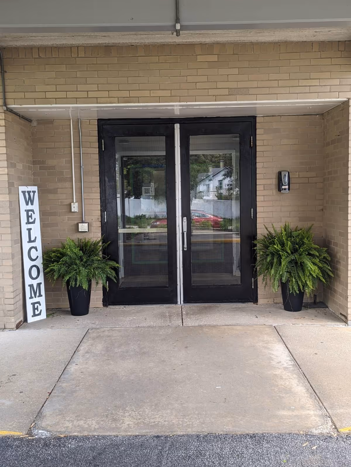 Double black framed glass doors set in a beige brick wall entrance with a vertical white sign reading 'WELCOME' on the left side and two green potted ferns on either side of the doors.