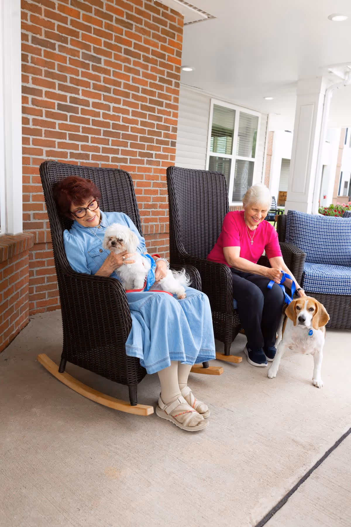 Two elderly women sitting on dark wicker rocking chairs on a covered porch. One woman, wearing a light blue dress and glasses, is holding a small white dog on her lap. The other woman, wearing a bright pink shirt and dark pants, is holding the leash of a beagle dog standing on the porch floor. The porch has a brick wall and white pillars, with windows and outdoor furniture in the background.