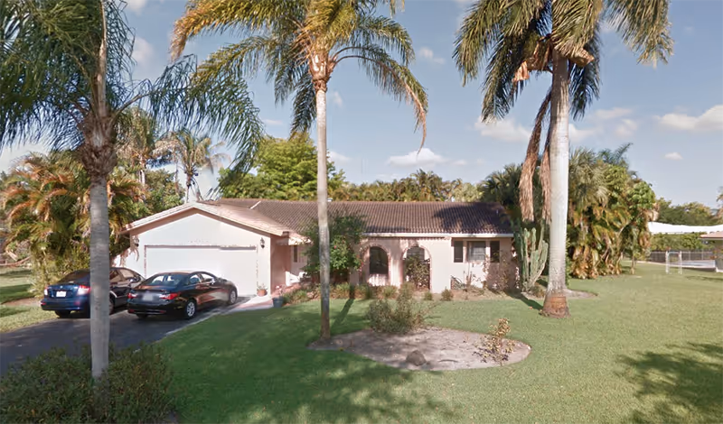 One-story pink senior facility building with a two-car driveway, palm trees, and manicured lawn.