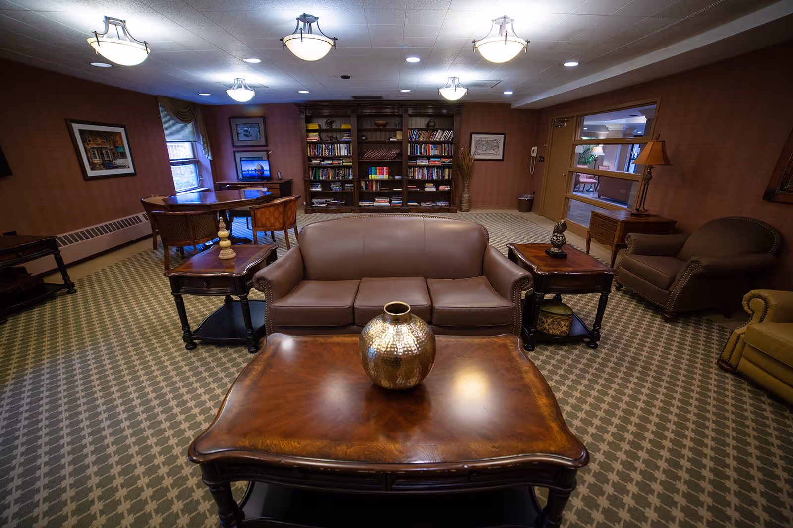 A cozy living room area in a senior living facility featuring a brown leather sofa, two armchairs, and a wooden coffee table with a decorative vase. Behind the sofa is a bookshelf filled with books, and to the left, a small table with chairs near a window. The room has patterned carpet flooring, warm lighting from ceiling fixtures, and framed artwork on the walls.