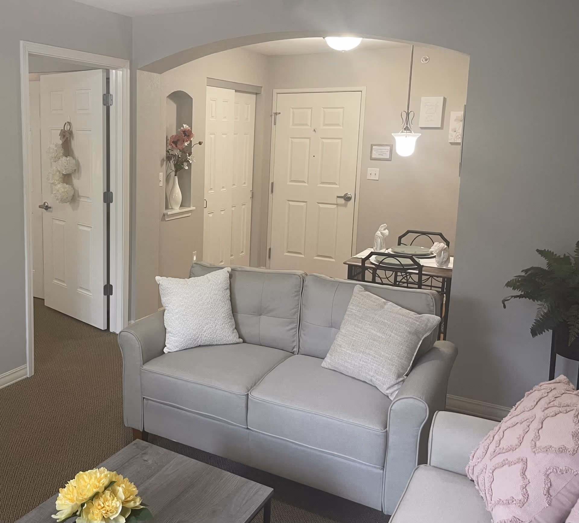 A cozy living room area with a light gray loveseat and a matching armchair with a pink textured pillow. A small wooden coffee table with a yellow flower arrangement is in front of the loveseat. In the background, there is a small dining table set for two with a hanging light fixture above it. The entry door and a closet with white doors are visible, along with a small wall niche holding a vase with flowers. The walls are painted light beige and the floor is carpeted.