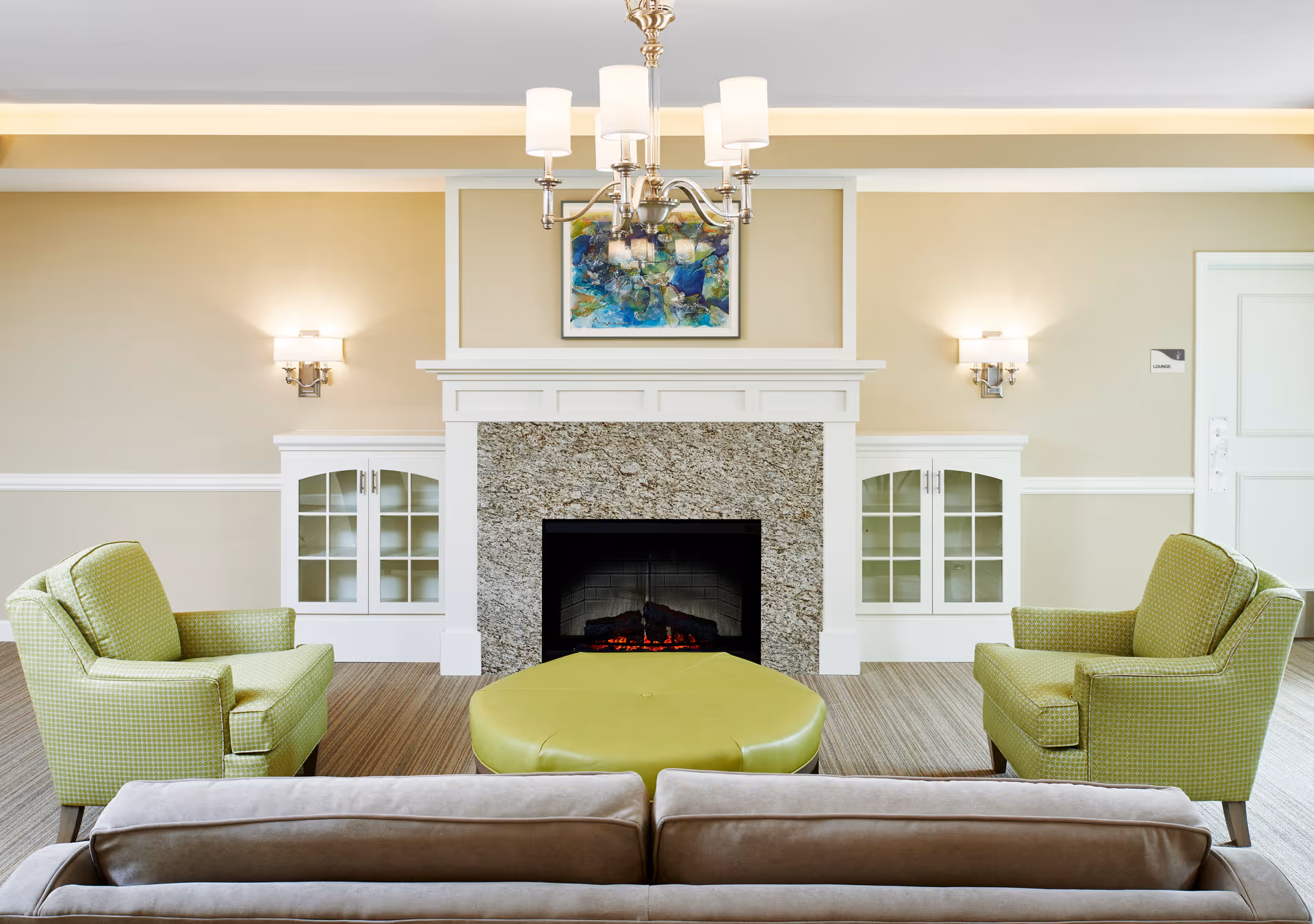 Well-lit living room with two green armchairs, a central green ottoman, a sofa in the foreground, and a fireplace with artwork above.