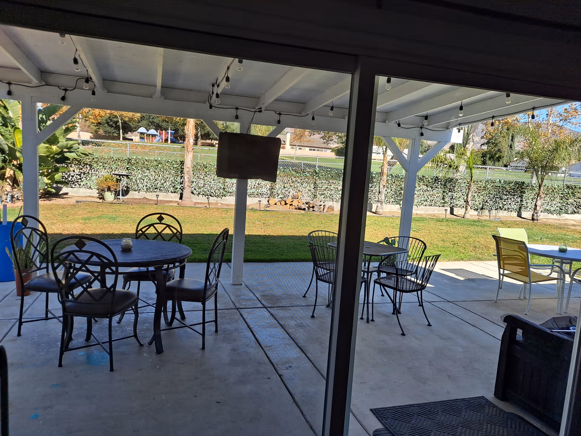 Covered patio with several metal tables and chairs overlooking a grassy backyard and a distant playground.