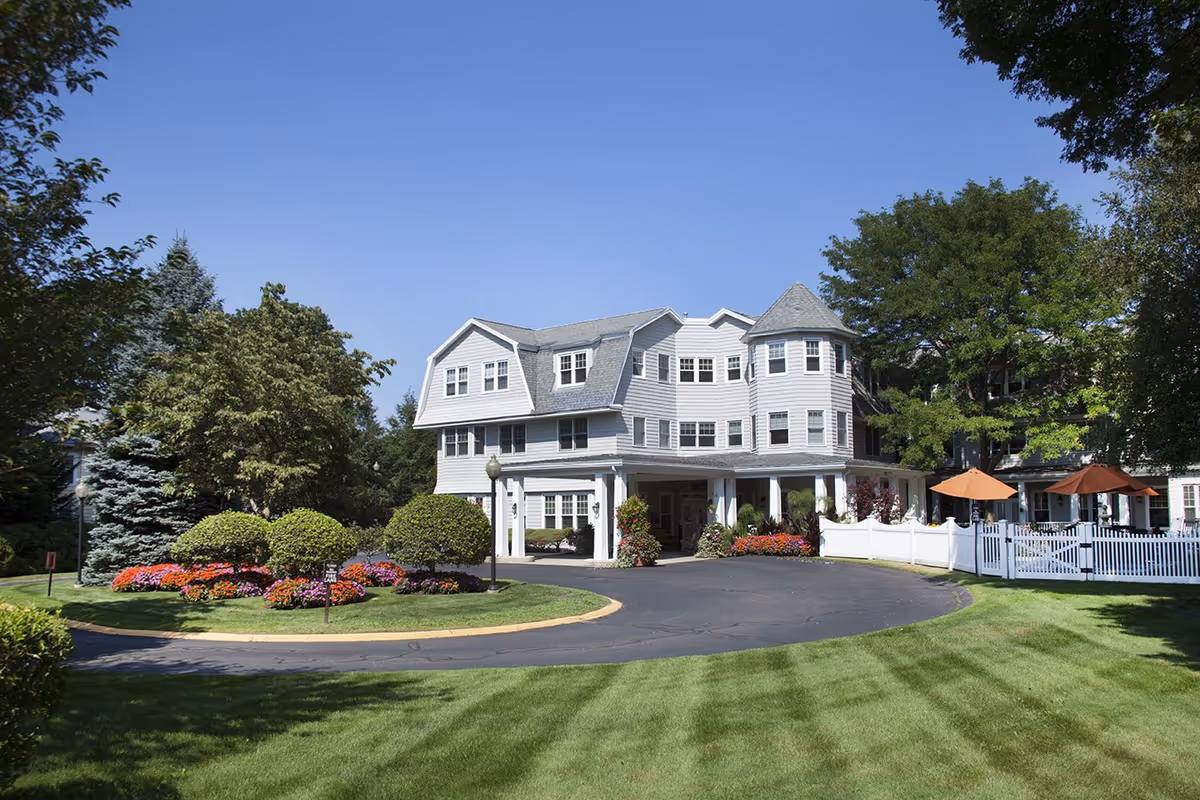 Three-story white building front with a circular driveway, manicured lawn, and colorful flower beds under a clear blue sky.