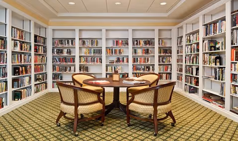 A cozy library room with white built-in bookshelves filled with books lining the walls. In the center, there is a round wooden table with four upholstered chairs around it. The carpet has a green and beige patterned design, and the ceiling has recessed lighting.