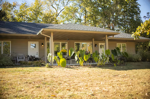 Single-story building with a covered porch, several windows and doors, surrounded by greenery and trees under a clear sky.