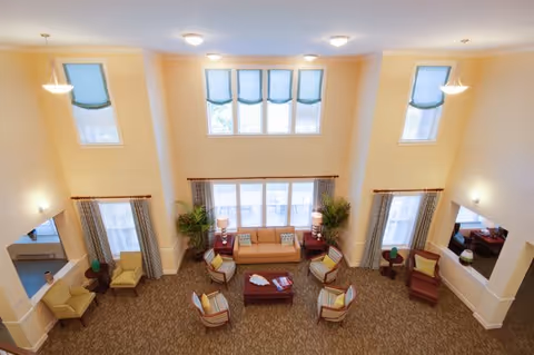 A spacious and well-lit living room area with high ceilings and multiple windows. The room features a central seating arrangement with a sofa, several armchairs, and a coffee table. There are side tables with lamps and potted plants near the windows. The walls are painted a soft yellow, and the carpet has a patterned design.
