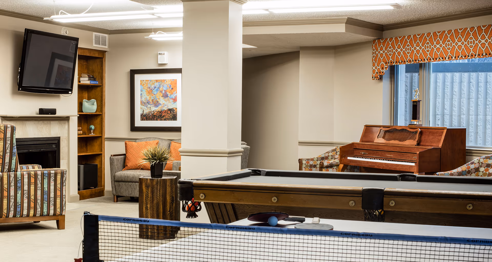 A cozy recreational room featuring a pool table and a ping pong table in the foreground. The room has a wall-mounted TV above a fireplace, a wooden bookshelf with decorative items, a gray sofa with orange pillows, and a wooden piano near a window with an orange patterned valance. Two patterned armchairs are placed near the piano.