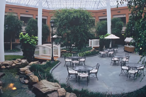 Indoor courtyard with a glass ceiling featuring several round metal tables and chairs with cushions, surrounded by lush green plants and trees. There is a small wooden bridge over a water feature with rocks, and a few umbrellas providing shade over some tables. The area is enclosed by a building with windows and doors.