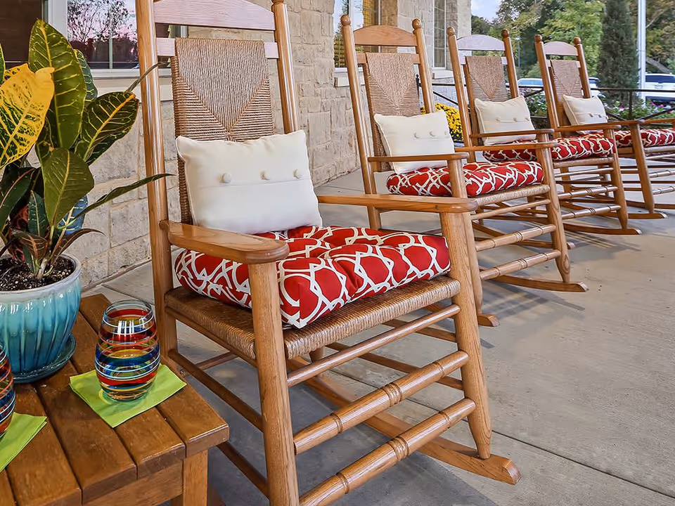 A row of wooden rocking chairs with red and white patterned seat cushions and white back pillows on a covered porch. A small wooden table with two colorful striped glasses and a potted plant is next to the chairs. The porch has a stone wall and windows in the background.