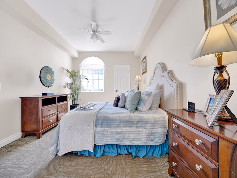 Sunlit bedroom with a neatly made bed, wooden nightstands and dresser, a lamp, and a potted plant by a window.