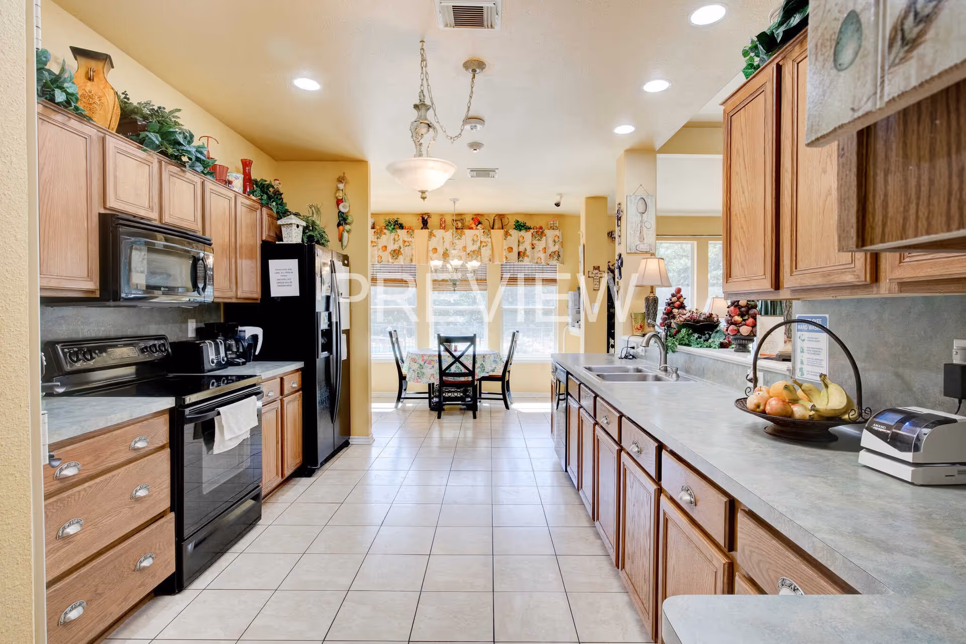 A bright and spacious kitchen with wooden cabinets on both sides, a black stove and microwave on the left, and a double sink on the right. The kitchen has a tiled floor and a dining area with a table and chairs near large windows at the far end. The countertops have various kitchen items and decorative plants above the cabinets.