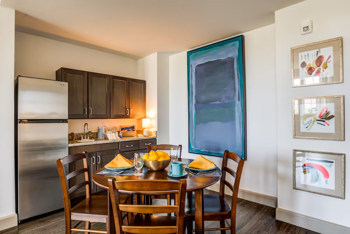 A dining area with a round wooden table set with four place settings, yellow napkins, glasses, and a bowl of lemons. Behind the table is a kitchenette with dark wooden cabinets, a stainless steel refrigerator, a sink, and a small lamp. The walls are decorated with a large abstract painting and three smaller framed abstract artworks.