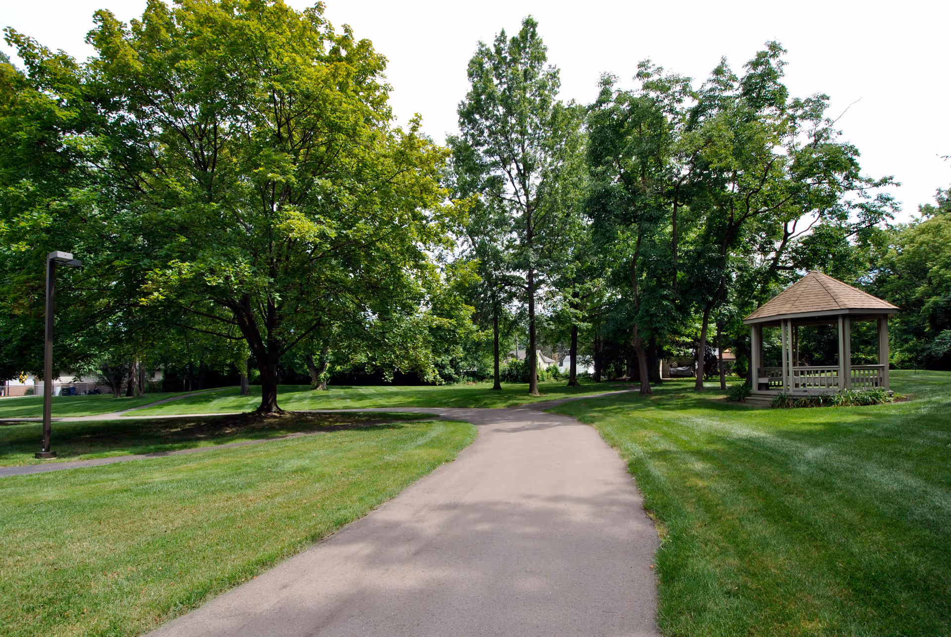 A paved walking path curves through a well-maintained grassy area with large green trees and a wooden gazebo on the right side under a bright sky.