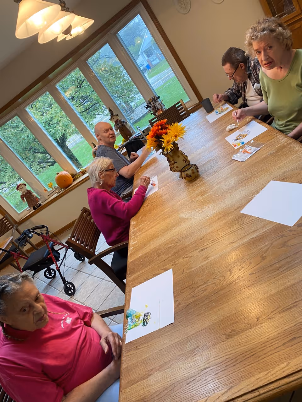 A group of elderly people sitting around a large wooden table in a bright room with large windows. They appear to be engaged in a painting or coloring activity, with papers and paint supplies on the table. There is a vase with colorful artificial flowers in the center of the table. Outside the windows, green trees and a house are visible. A walker is positioned near one of the chairs.