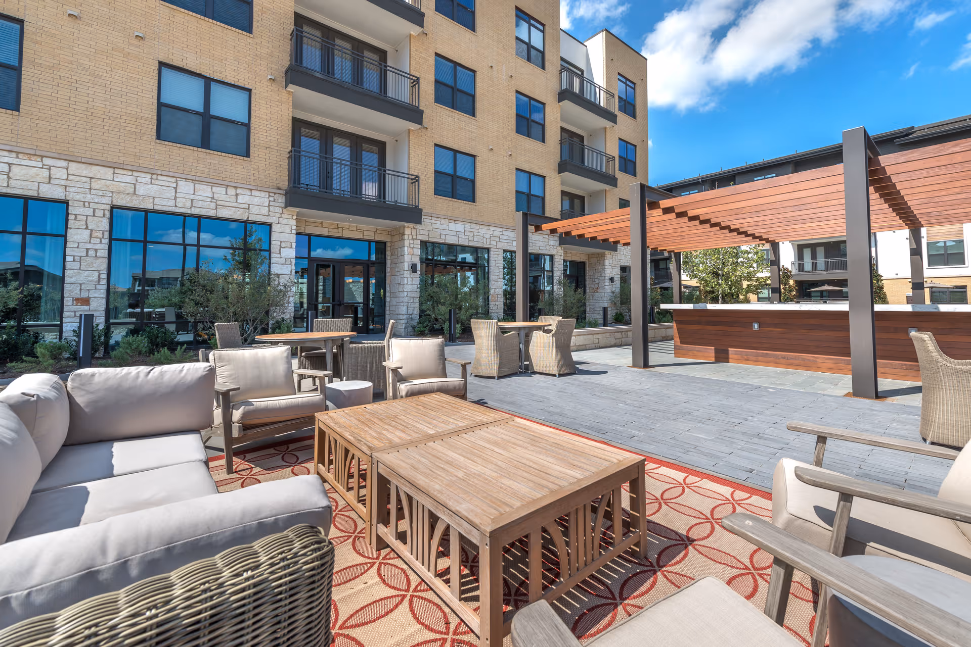 Outdoor patio area at Overture Fairview featuring cushioned wicker seating, wooden tables, a patterned outdoor rug, and a wooden pergola structure with a clear blue sky above. The building exterior has large windows and balconies.