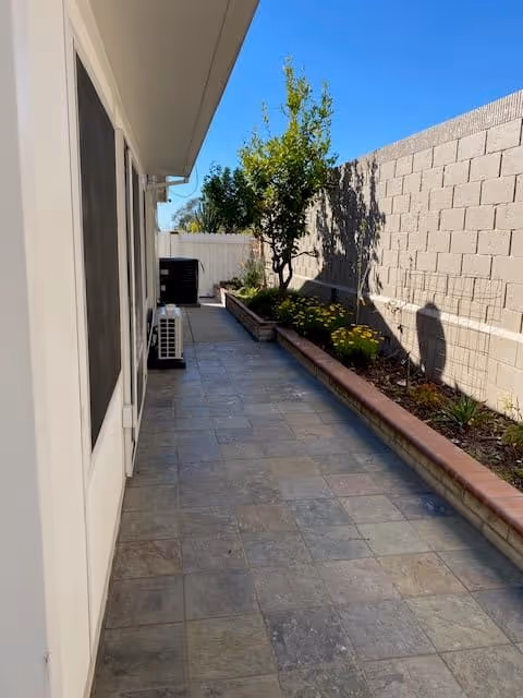 A narrow outdoor patio area with tiled flooring, a raised garden bed with plants and a small tree along a brick wall, and the side of a building with windows and air conditioning units under a clear blue sky.