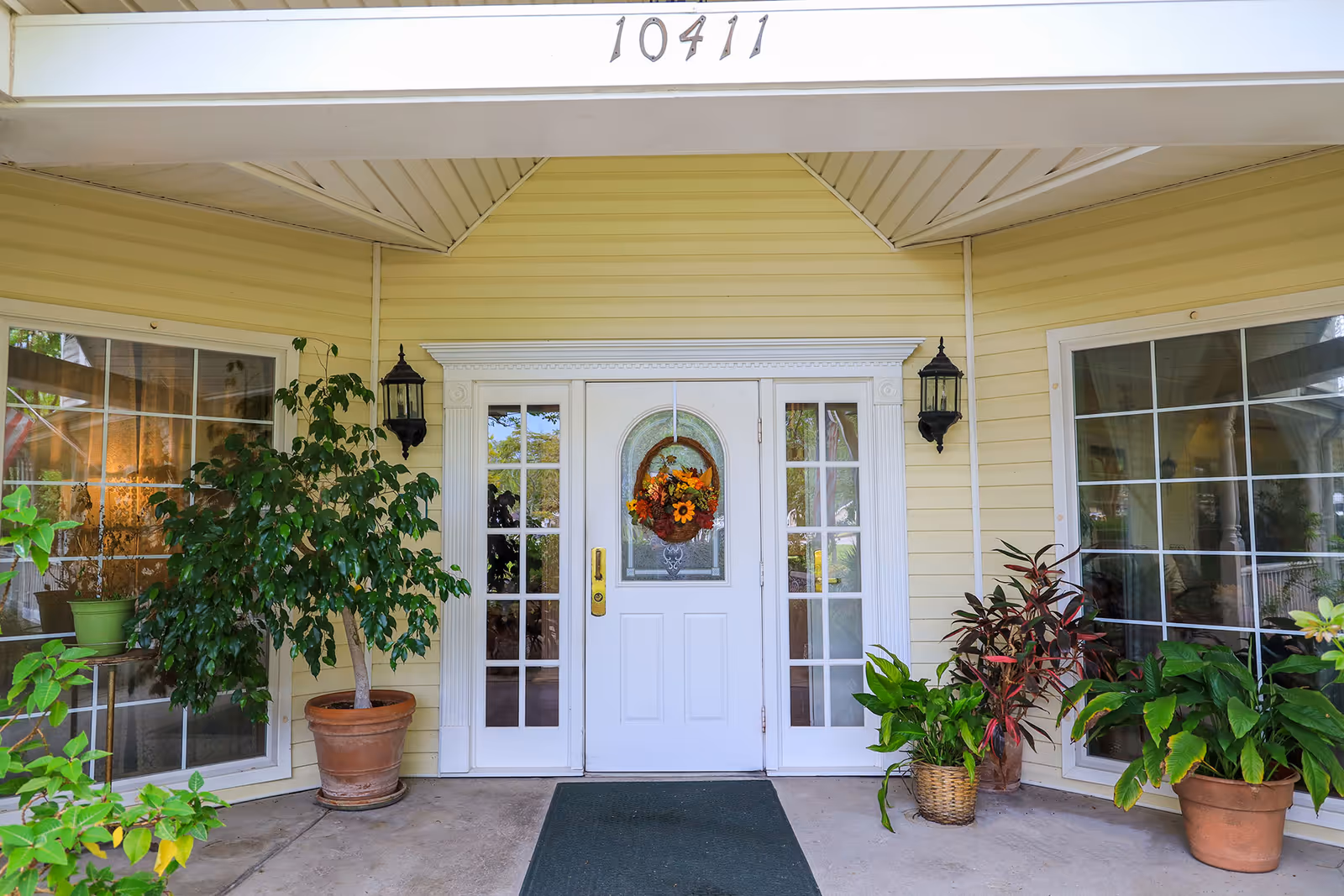 Front entrance of a yellow-sided building with white double doors decorated with a wreath, flanked by sidelights, large windows and potted plants, with the address number 10411 above.