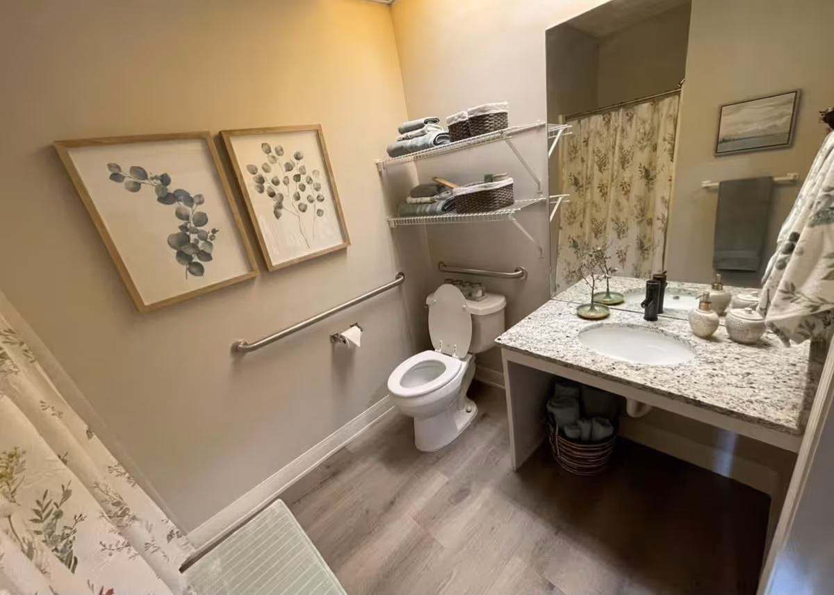 A clean and well-organized bathroom with a toilet, a granite countertop sink, and a shower with a floral curtain. Two framed botanical prints hang on the wall above a grab bar. Shelves above the toilet hold neatly folded towels and baskets. A towel rack with a green towel is mounted on the wall next to the sink.