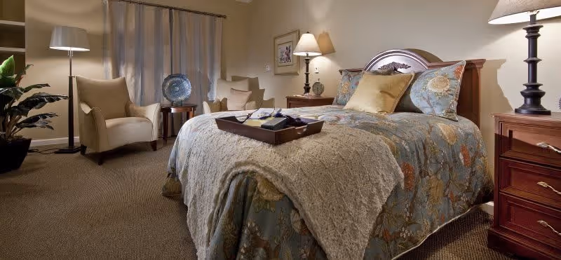 A cozy bedroom with a large bed covered in a floral-patterned comforter and a beige knitted throw. There are two wooden nightstands with lamps on either side of the bed. In the background, two beige armchairs are positioned near a window with curtains, and a small round table with a decorative plate is placed between them. The room has warm lighting and a carpeted floor.