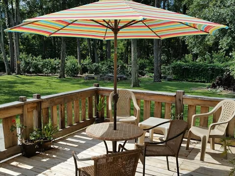 Wooden deck with a round table and four chairs, shaded by a large striped umbrella. The deck overlooks a green lawn with trees and bushes in the background.