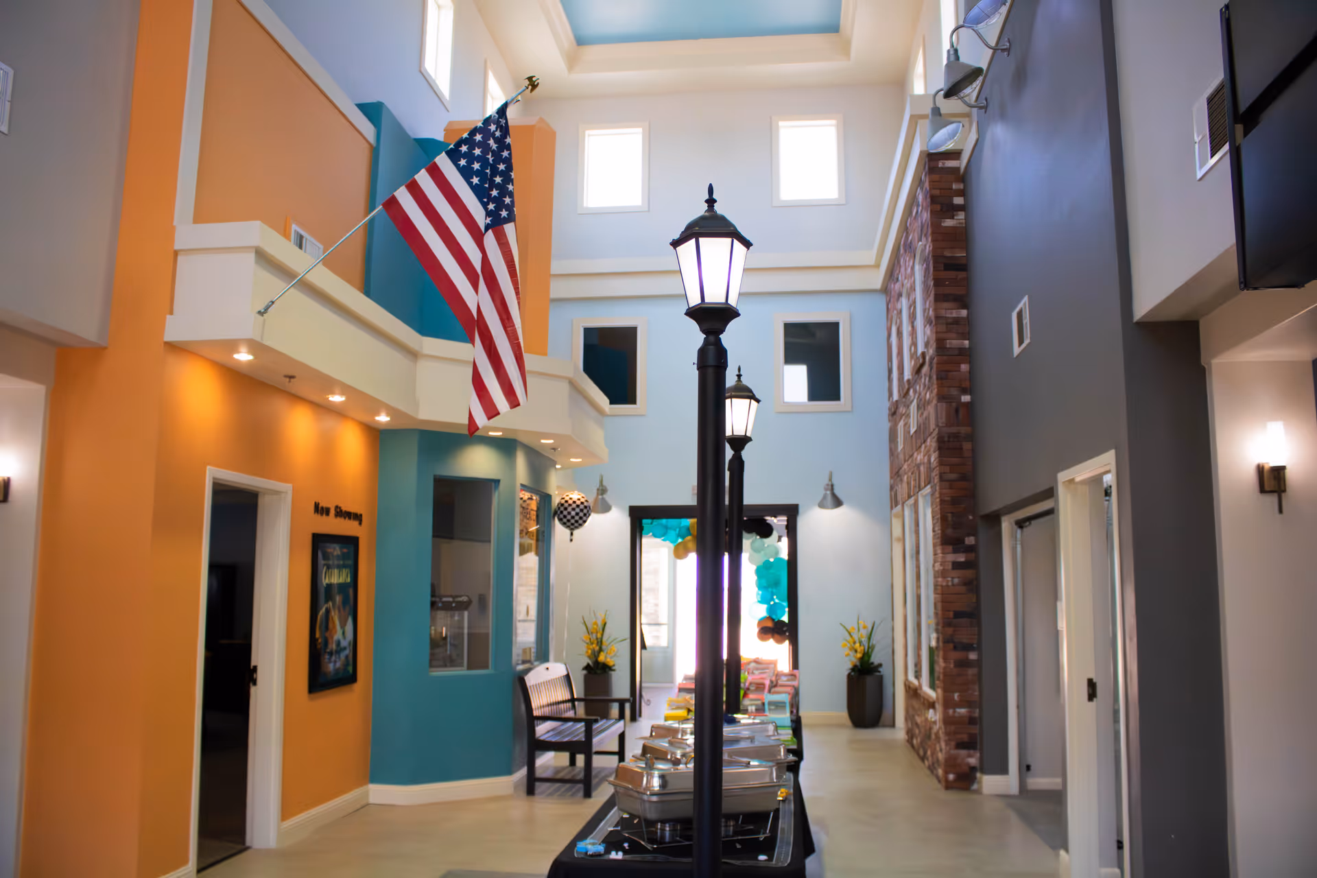 Bright two-story interior lobby with an American flag, streetlamp-style lights, colorful walls, seating and a buffet table under a skylight.