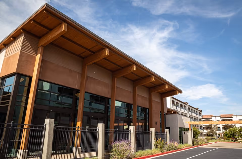 Exterior view of a modern building with large windows and wooden beams under a blue sky with some clouds. The building is surrounded by a black metal fence and landscaped with plants and flowers. A parking area is visible in the foreground.