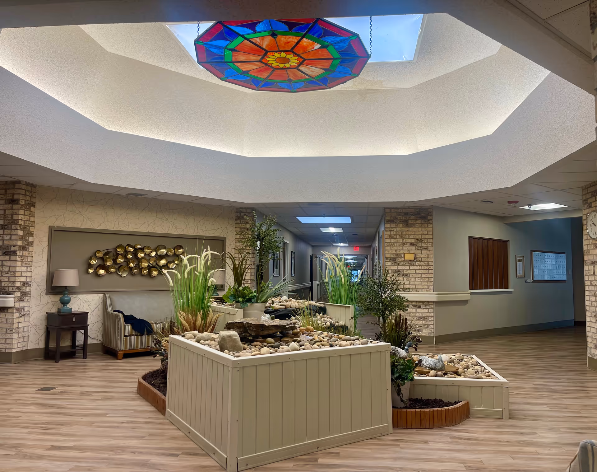 Interior view of a senior living facility hallway with a central indoor garden featuring plants and rocks. The ceiling has a skylight with a colorful stained glass decoration. There is a seating area with a striped armchair and a side table with a lamp on the left side. The walls are decorated with brick accents and artwork.