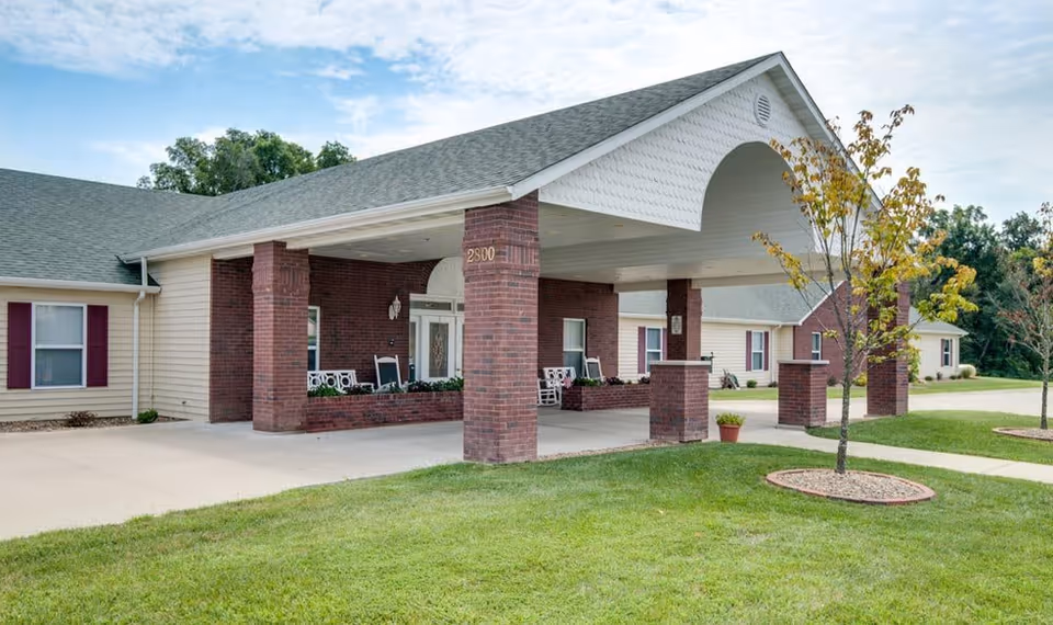 Exterior view of a senior living facility with a covered entrance supported by brick columns. The building has beige siding, a gray shingled roof, and several windows with maroon shutters. There is a small tree and green lawn in the foreground under a partly cloudy sky.