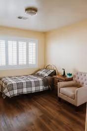 Simple furnished bedroom with a single bed, nightstand, armchair, and shuttered window.