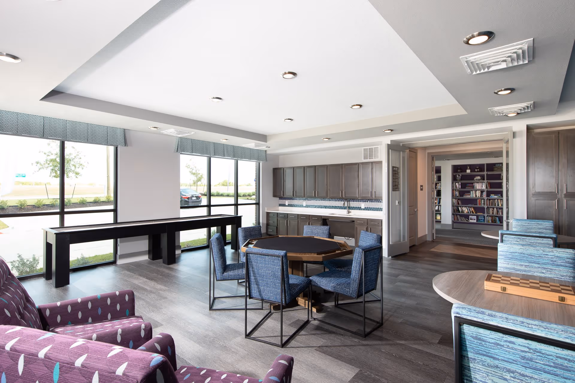 A bright and modern common area with large windows letting in natural light. The room features a round table with six blue upholstered chairs, a purple armchair with a patterned design, and a long black shuffleboard table near the windows. There are wooden cabinets and a countertop with a sink along one wall. In the background, an open doorway leads to a room with bookshelves filled with books.