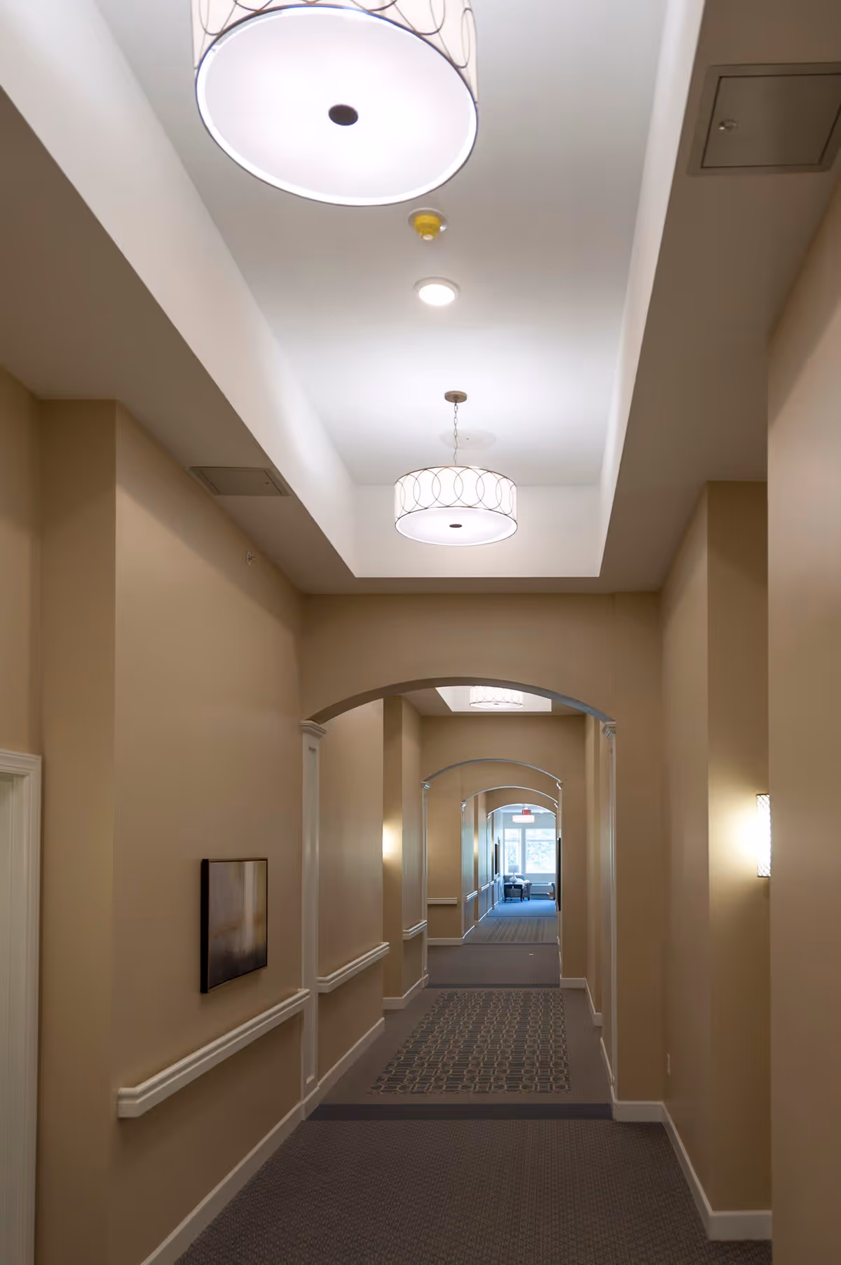 A well-lit hallway with beige walls and carpeted floor featuring patterned runners. The ceiling has recessed lighting and two large circular pendant lights. The hallway has several archways and handrails along the walls, with a framed picture on the left wall and a window visible at the far end.