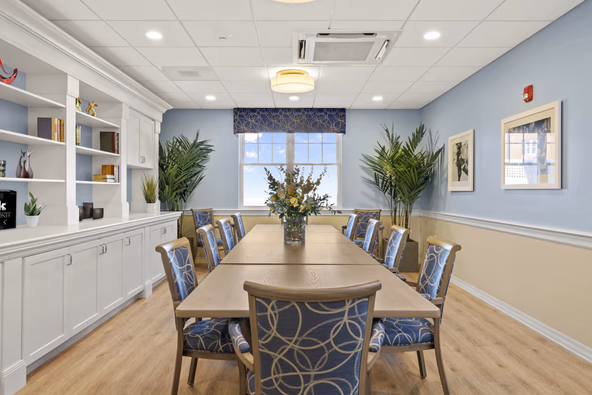 A well-lit dining room with a long wooden table surrounded by eight chairs upholstered in blue fabric with circular patterns. A vase with flowers is centered on the table. The room has light blue walls with white trim, two large potted plants near the window, built-in white cabinetry with shelves holding books and decorative items, and two framed pictures on the right wall. A window with a blue valance lets in natural light.