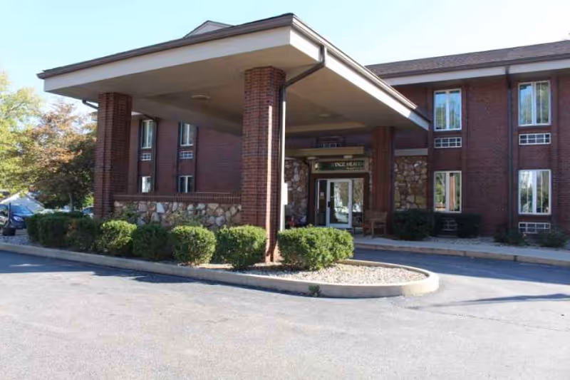 Exterior view of The Grove Health and Rehab building entrance with a covered drop-off area supported by brick columns, surrounded by bushes and a paved driveway.