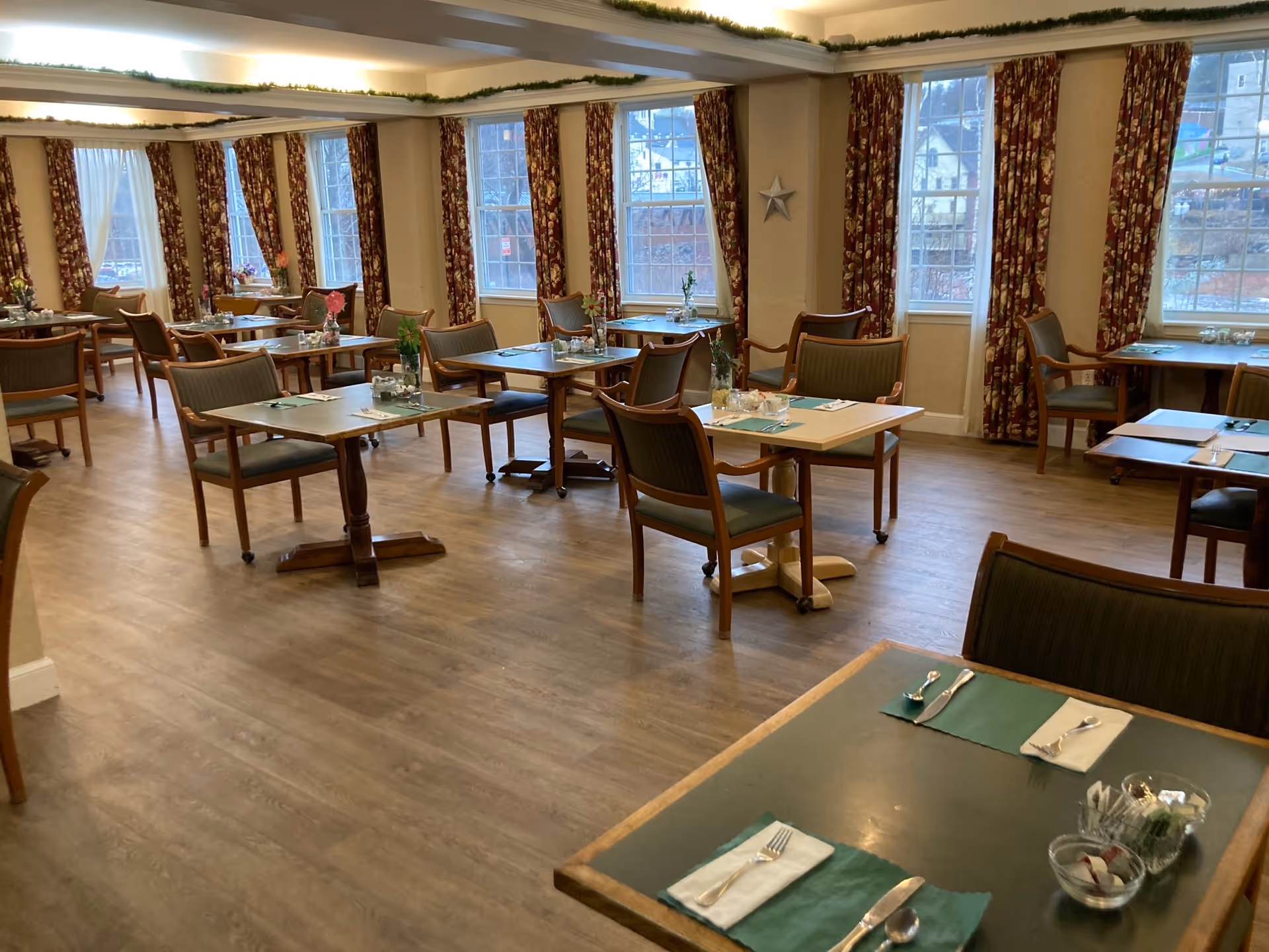 Dining room with several set tables and chairs, large windows with floral curtains, and wood-look flooring.
