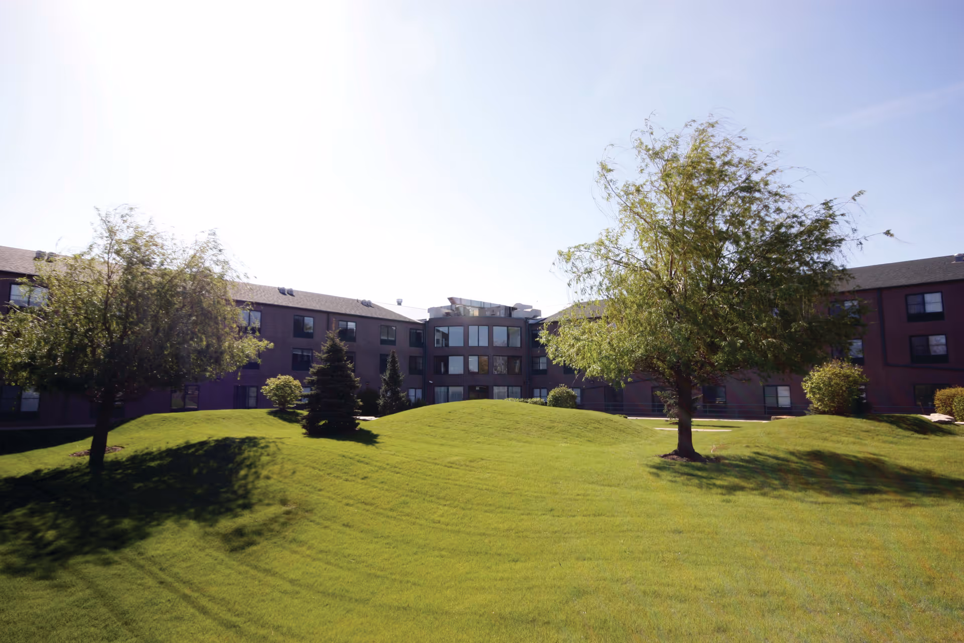 Exterior view of a senior living facility building with a well-maintained green lawn and several trees under a clear sky.