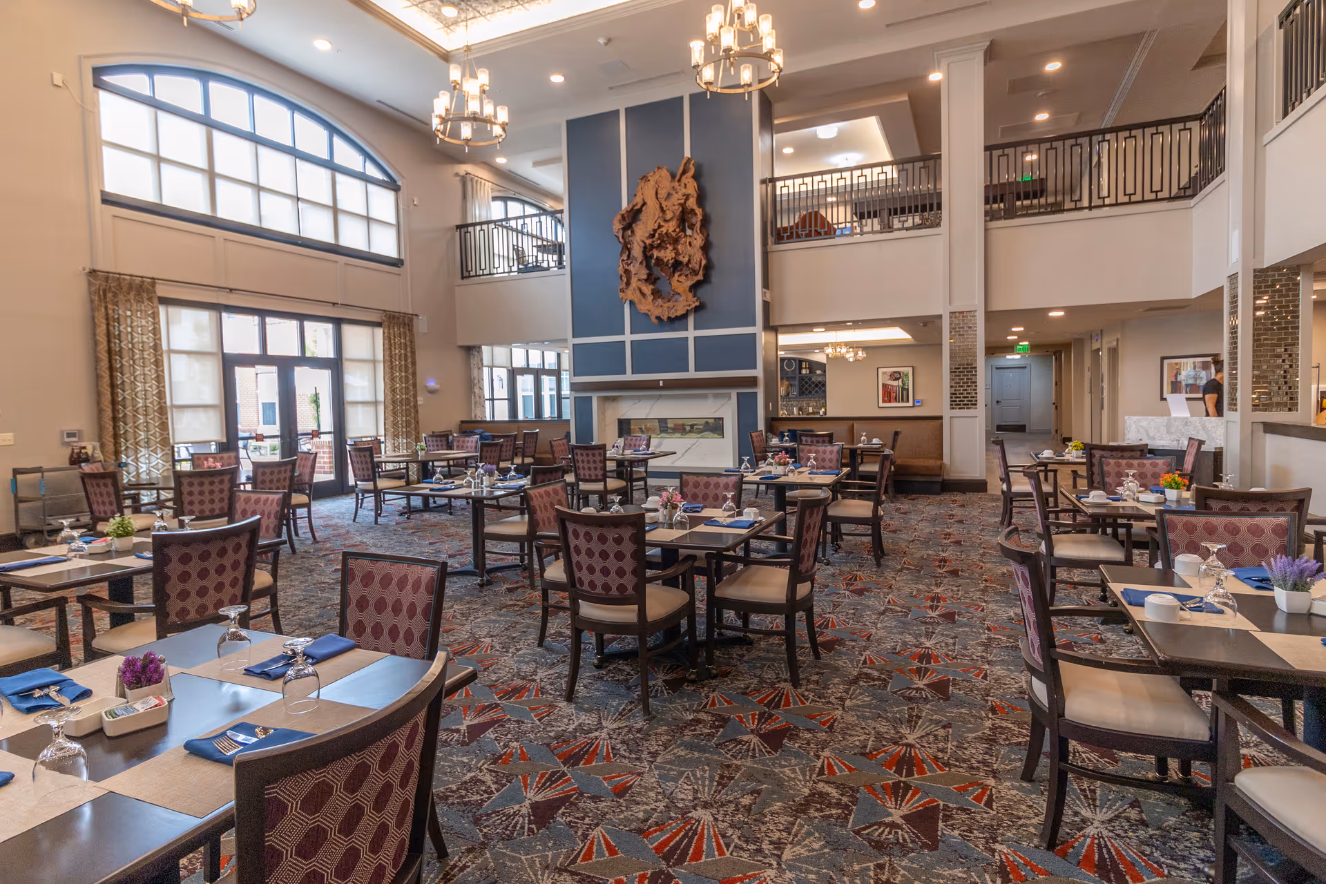 Spacious two-story dining room with multiple set tables, patterned carpet, large windows, and a decorative wall sculpture.
