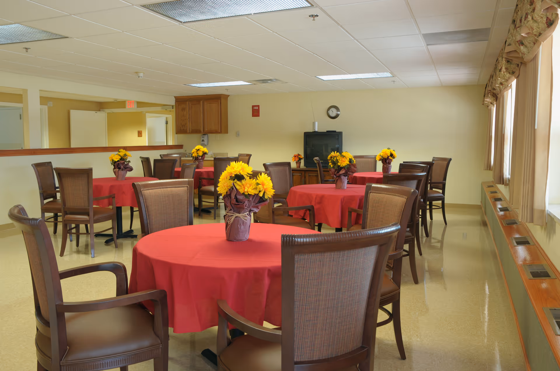 A dining room with several round tables covered with red tablecloths, each decorated with a vase of yellow flowers. The room has wooden chairs around the tables, large windows with curtains on the right side, and a TV on a stand against the far wall.