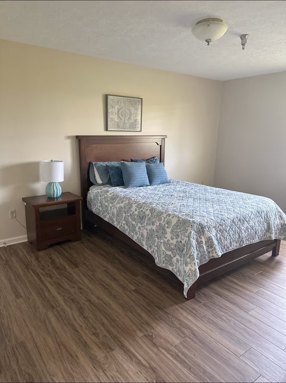 A bedroom with a wooden bed frame and a bed made with a blue and white patterned quilt and several blue pillows. There is a wooden nightstand with a blue lamp on it next to the bed. The room has light-colored walls, wood flooring, and a framed artwork hanging above the bed.