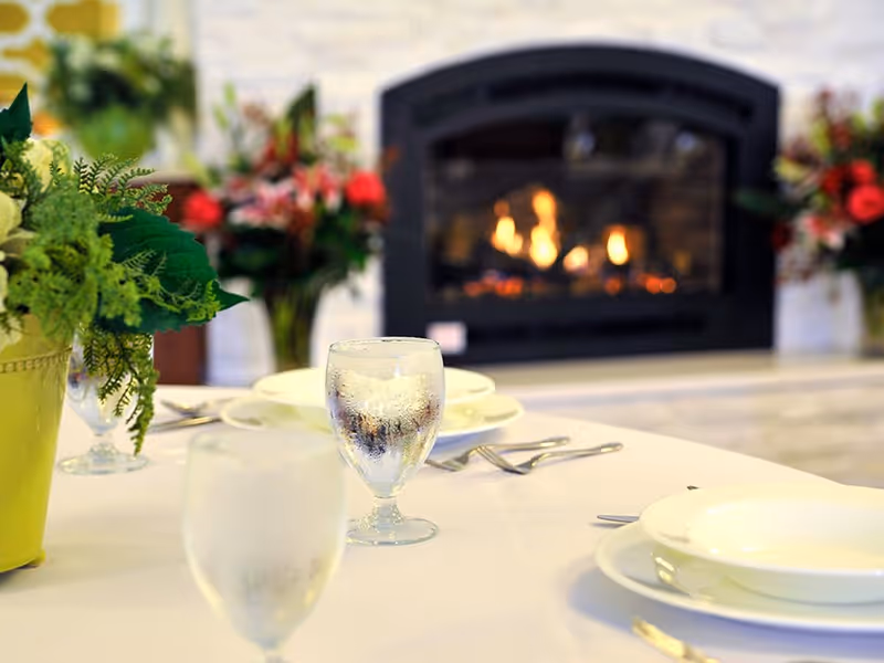 A dining table set with water glasses, plates and floral centerpieces placed in front of a lit fireplace.