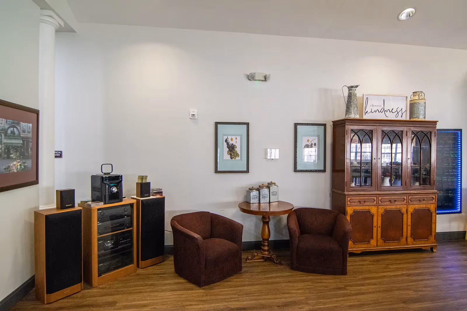 A cozy interior corner of a senior living facility featuring two brown upholstered armchairs separated by a round wooden table with decorative jars on top. Behind the chairs, there are two framed botanical prints on the wall. To the right, there is a wooden cabinet with glass doors displaying items inside, topped with decorative pieces and a sign that reads 'choose kindness.' On the left side, there is a stereo system with speakers on a wooden stand. The floor is wood, and the walls are painted light gray.