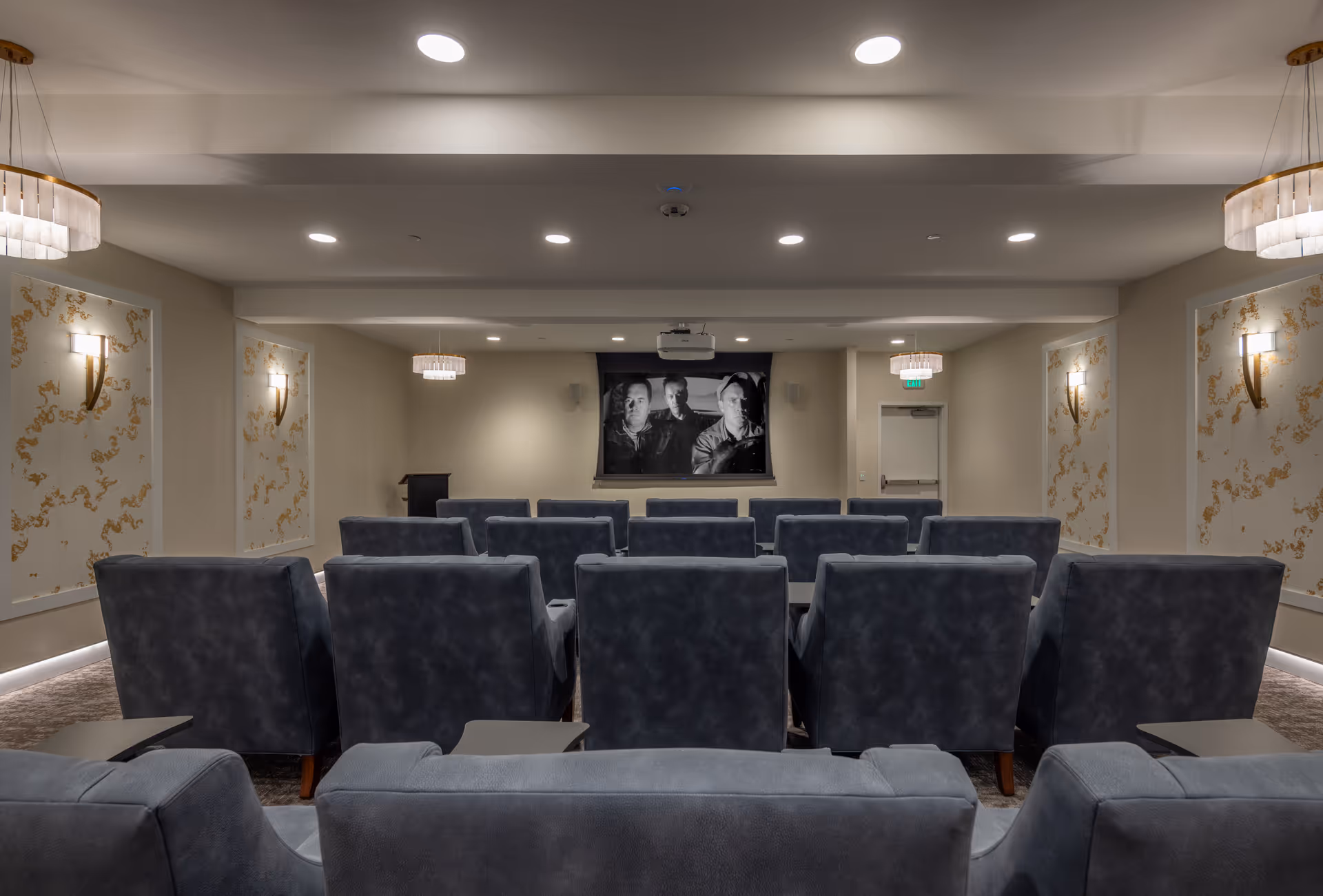 A small theater room with multiple rows of gray upholstered chairs facing a screen showing a black and white movie. The room has soft lighting with wall sconces and ceiling lights, and decorative wall panels with gold accents.