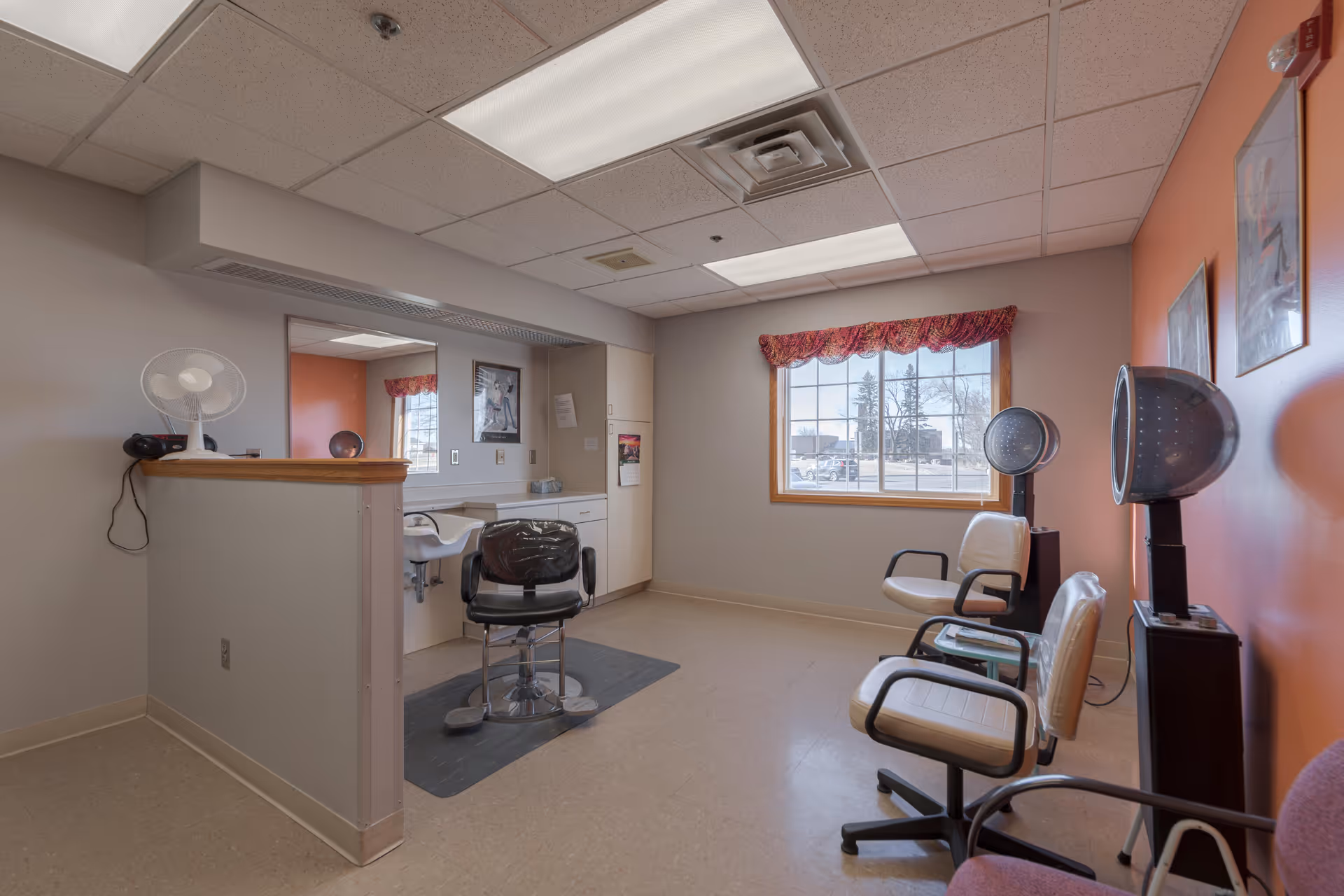 Interior view of a hair salon area in a senior living facility with a styling chair in front of a large mirror, a small fan on a half wall, and three waiting chairs with hair dryers near a window with a red valance.